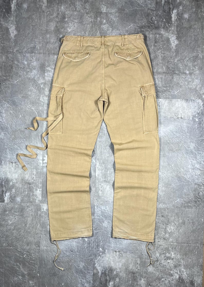 May include: Tan cargo pants featuring two side cargo pockets, two back pockets with flaps, and a waistband with belt loops. The pants include long fabric ties attached to the left cargo pocket and adjustable drawstrings at the bottom hems.