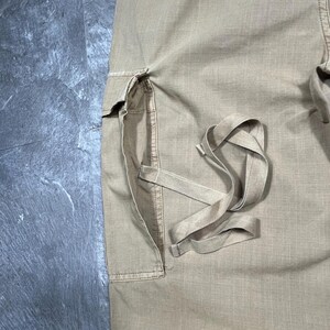 May include: Tan cargo pants featuring a large side pocket with a flap closure and long, dangling fabric straps attached to the pocket area. The fabric has a visible woven texture, and the design includes reinforced stitching along the pocket seams.