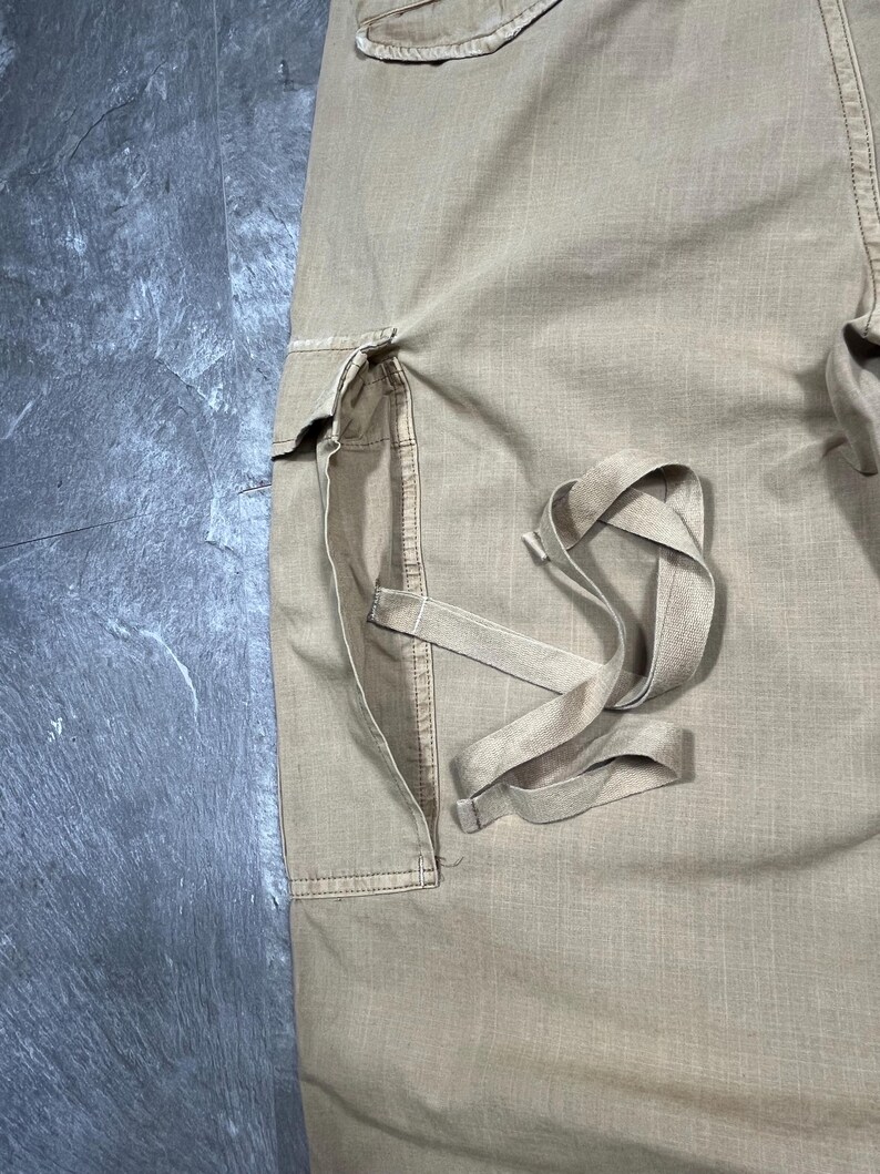 May include: Tan cargo pants featuring a large side pocket with a flap closure and long, dangling fabric straps attached to the pocket area. The fabric has a visible woven texture, and the design includes reinforced stitching along the pocket seams.