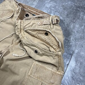 May include: Tan cargo pants featuring multiple pockets with button closures, a drawstring waist, and reinforced stitching. The fabric has a distressed, worn-in appearance with visible frayed edges along the pocket flaps and waistband seams.
