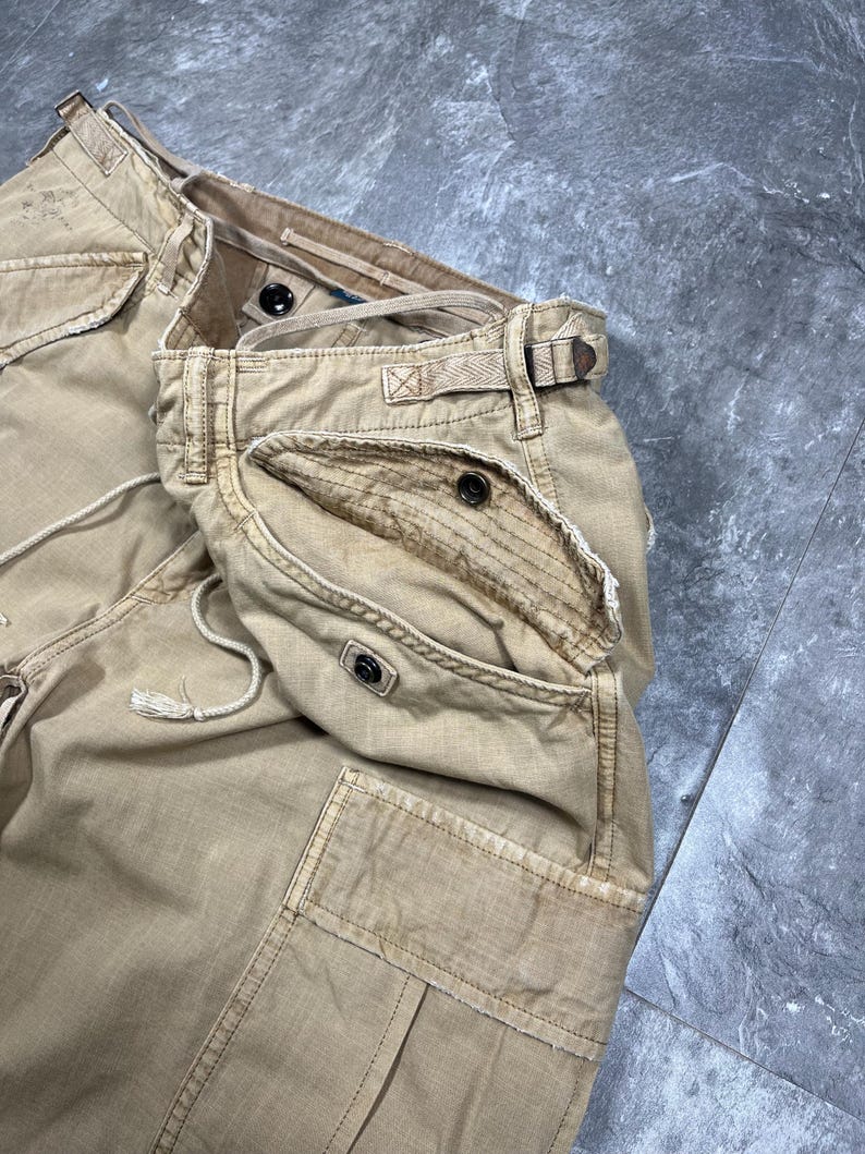 May include: Tan cargo pants featuring multiple pockets with button closures, a drawstring waist, and reinforced stitching. The fabric has a distressed, worn-in appearance with visible frayed edges along the pocket flaps and waistband seams.