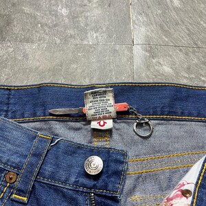 May include: Close-up of blue denim jeans with a button closure and a zipper. The jeans have yellow stitching and a True Religion brand tag. A small knife-shaped keychain is attached to the jeans.