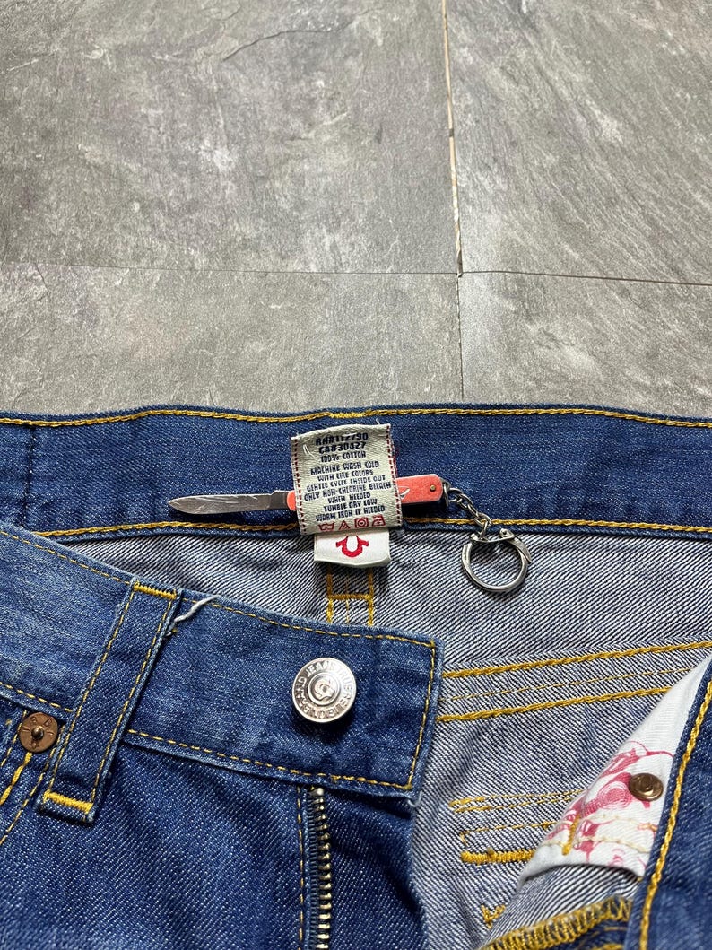 May include: Close-up of blue denim jeans with a button closure and a zipper. The jeans have yellow stitching and a True Religion brand tag. A small knife-shaped keychain is attached to the jeans.