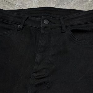 Op de afbeelding: Close-up van een zwarte spijkerbroek. De jeans heeft een knoopsluiting, riemlussen en zakken. De stof lijkt licht beschadigd, met een kleine scheur zichtbaar op de broekspijp. De algehele stijl is casual en modern.