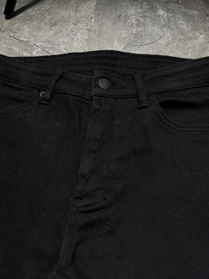 Op de afbeelding: Close-up van een zwarte spijkerbroek. De jeans heeft een knoopsluiting, riemlussen en zakken. De stof lijkt licht beschadigd, met een kleine scheur zichtbaar op de broekspijp. De algehele stijl is casual en modern.