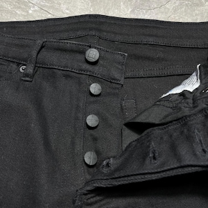 Op de afbeelding: Close-up van een zwarte spijkerbroek. De jeans heeft een knoopsluiting met vier donkergrijze knopen en een knoopsluiting in de tailleband. De stof lijkt een stevige, donker gewassen denim te zijn.