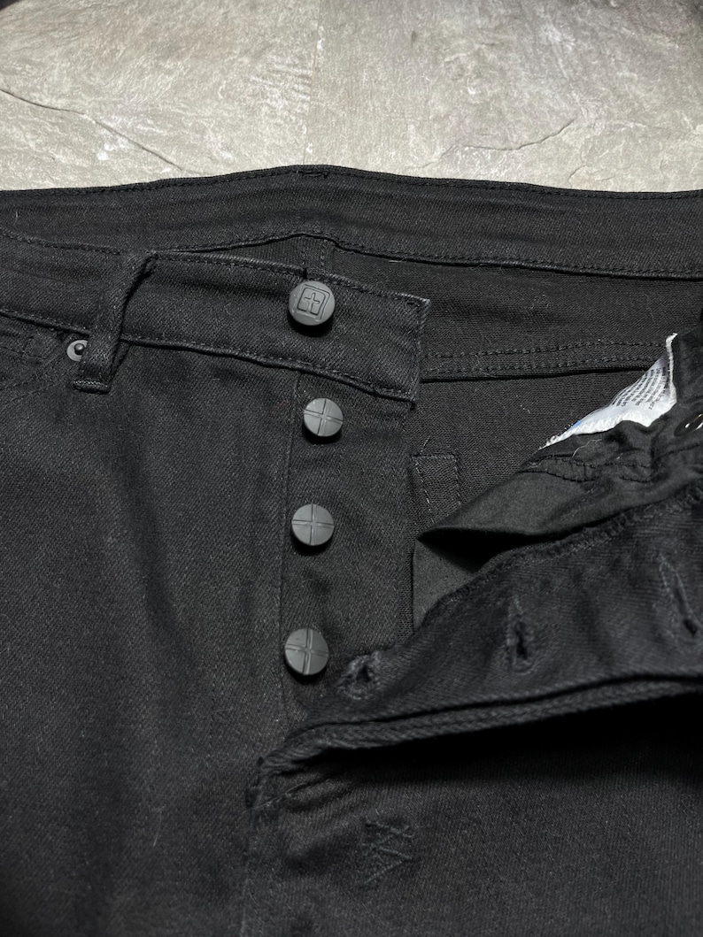 Op de afbeelding: Close-up van een zwarte spijkerbroek. De jeans heeft een knoopsluiting met vier donkergrijze knopen en een knoopsluiting in de tailleband. De stof lijkt een stevige, donker gewassen denim te zijn.