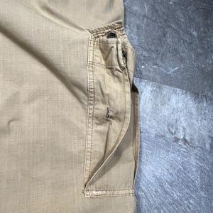 May include: A pair of tan cargo pants featuring a large side pocket with a flap closure, reinforced stitching, and a metal snap button. The fabric has a visible grid-like texture, and the pocket includes a vertical slit opening with frayed edges.