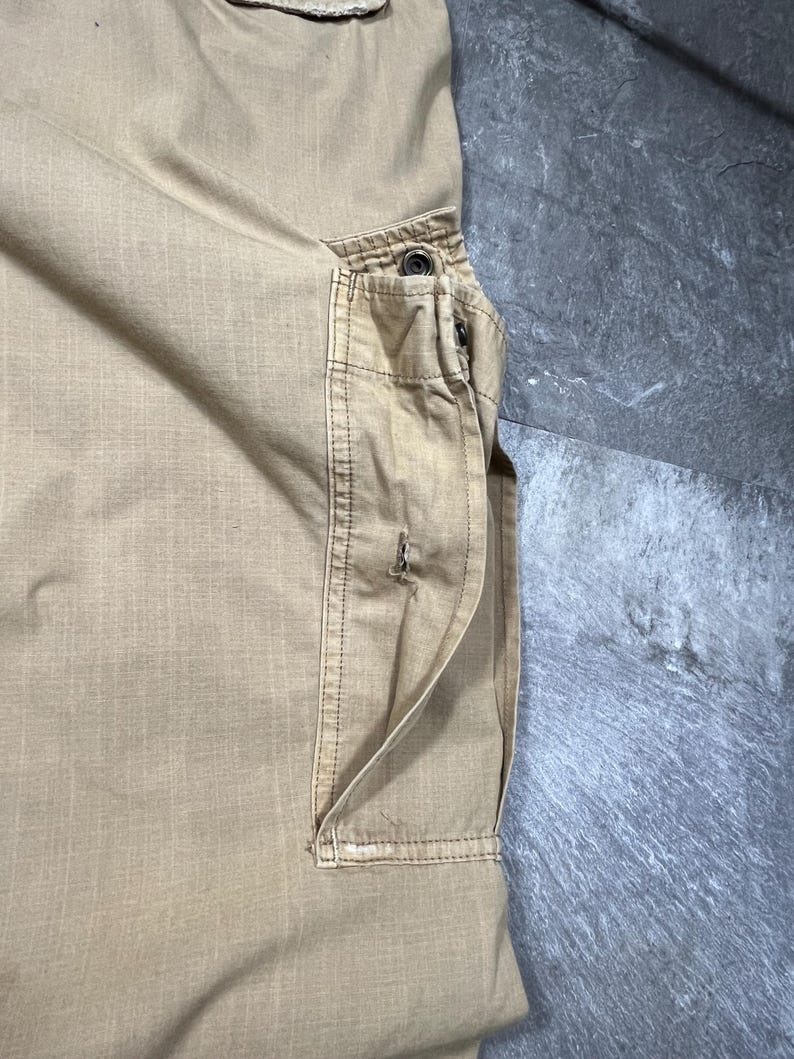 May include: A pair of tan cargo pants featuring a large side pocket with a flap closure, reinforced stitching, and a metal snap button. The fabric has a visible grid-like texture, and the pocket includes a vertical slit opening with frayed edges.