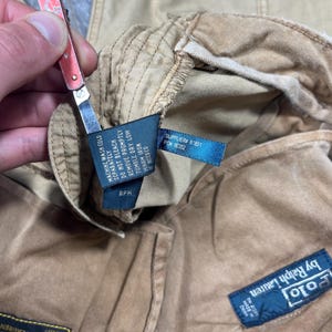 May include: Tan corduroy pants featuring a blue Polo by Ralph Lauren label, a black care tag with washing instructions, and a rectangular patch with text including "PANT, MANS" and "POLO RALPH LAUREN" visible on the interior fabric.