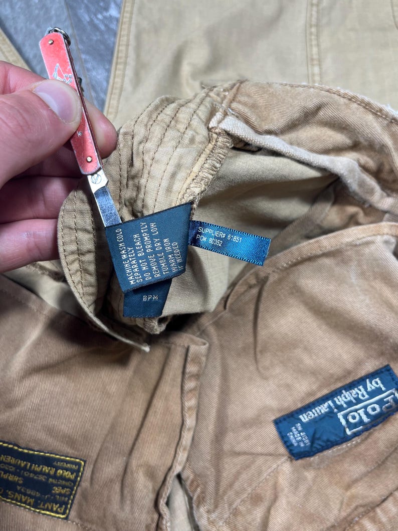 May include: Tan corduroy pants featuring a blue Polo by Ralph Lauren label, a black care tag with washing instructions, and a rectangular patch with text including "PANT, MANS" and "POLO RALPH LAUREN" visible on the interior fabric.