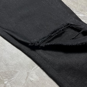 Op de afbeelding: Close-up van een zwarte denim jeans met een gerafelde, gerafelde zoom. De stof is een donkere, effen kleur, met zichtbare textuur. De jeans is opgevouwen en toont de rand van de zoom.