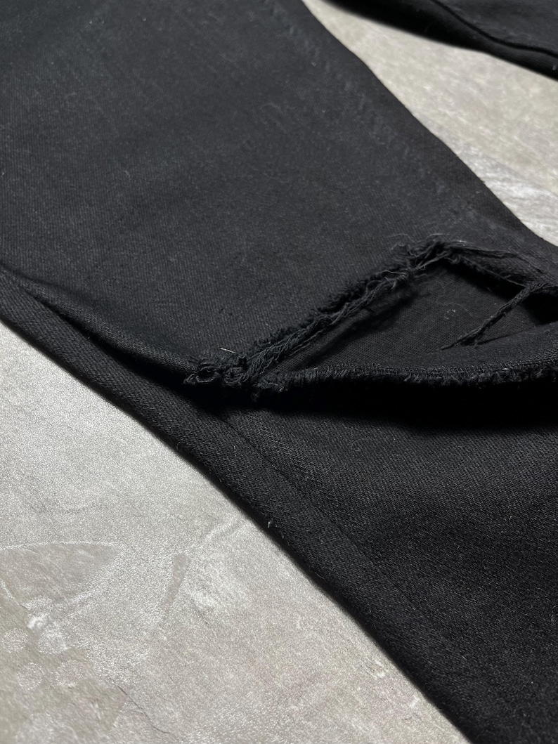 Op de afbeelding: Close-up van een zwarte denim jeans met een gerafelde, gerafelde zoom. De stof is een donkere, effen kleur, met zichtbare textuur. De jeans is opgevouwen en toont de rand van de zoom.
