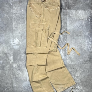 May include: Tan cargo pants featuring a large side pocket with a flap, a rear pocket with a flap, and adjustable fabric ties attached to the side. The pants have a structured waistband with belt loops and drawstrings at the bottom of the pant legs.