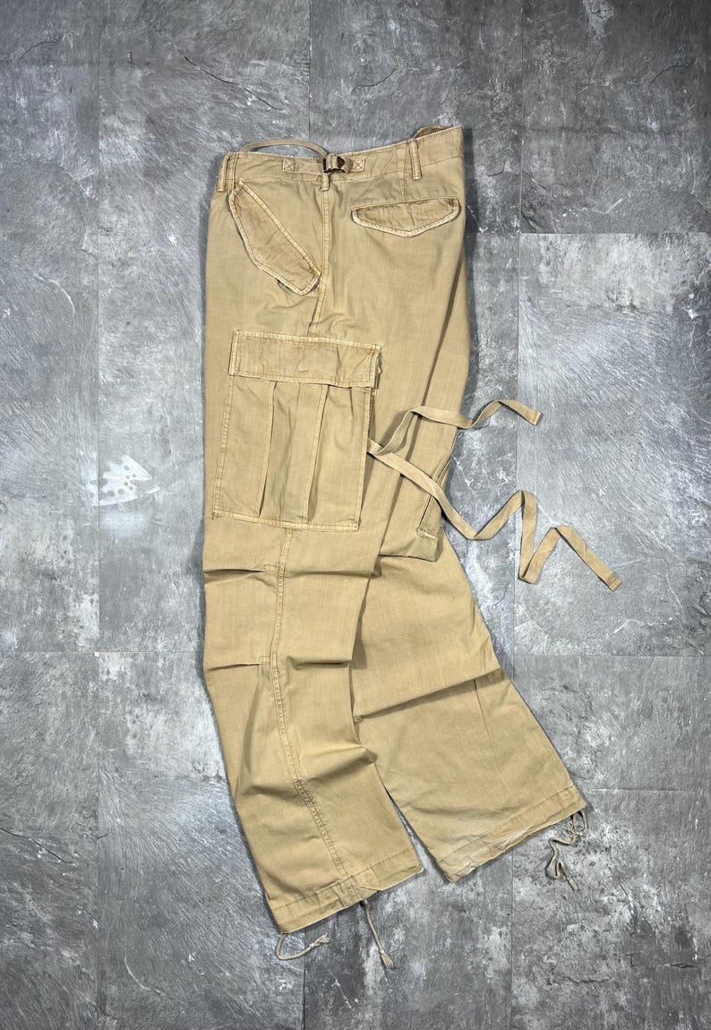 May include: Tan cargo pants featuring a large side pocket with a flap, a rear pocket with a flap, and adjustable fabric ties attached to the side. The pants have a structured waistband with belt loops and drawstrings at the bottom of the pant legs.