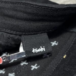 Op de afbeelding: Close-up van een zwarte spijkerbroek met de merknaam "+subi" in witte letters op een zwart label. De jeans heeft witte stervormige stiksels aan de binnenkant.