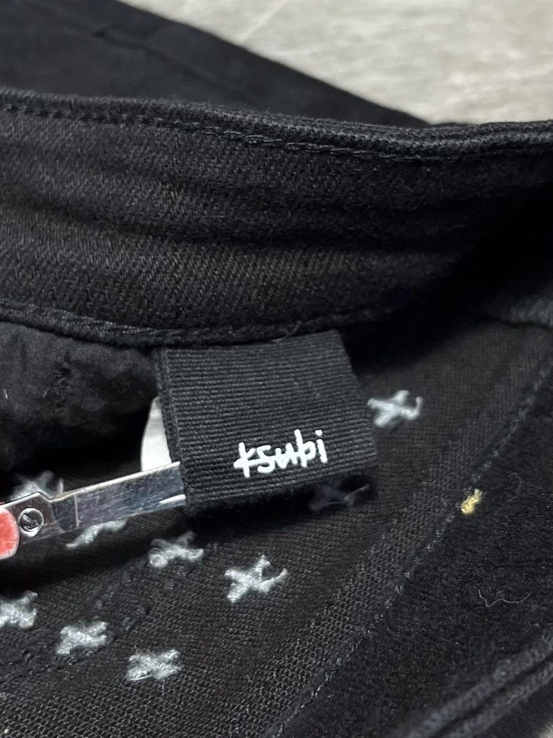 Op de afbeelding: Close-up van een zwarte spijkerbroek met de merknaam "+subi" in witte letters op een zwart label. De jeans heeft witte stervormige stiksels aan de binnenkant.