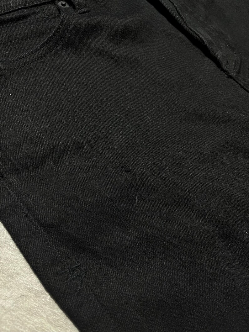 Op de afbeelding: Close-up van een zwarte spijkerbroek. De jeans heeft een klassiek ontwerp met vijf zakken, met een zichtbare voorzak en een klein logodetail aan de onderkant. De stof lijkt een stevige, donker gewassen denim te zijn.