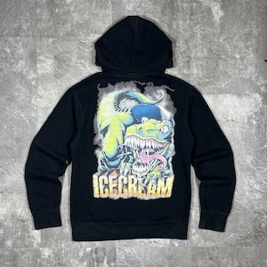 Puede incluir: Una sudadera negra con capucha que presenta un gráfico en la espalda de un dinosaurio verde con una gorra de béisbol azul, con la boca abierta y la lengua fuera. Debajo del dinosaurio, la palabra ICECREAM está impresa en letras amarillas.