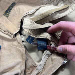 May include: Tan cotton pants featuring a button fly, belt loops, and a black interior label that reads 34/34 and 100% cotton care instructions over. The garment includes a rectangular black brand patch with yellow text detailing Polo Ralph Lauren.