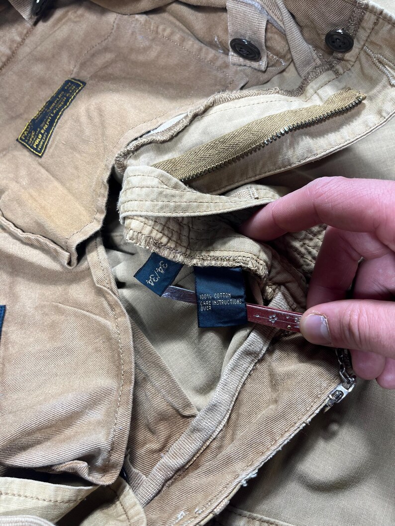 May include: Tan cotton pants featuring a button fly, belt loops, and a black interior label that reads 34/34 and 100% cotton care instructions over. The garment includes a rectangular black brand patch with yellow text detailing Polo Ralph Lauren.