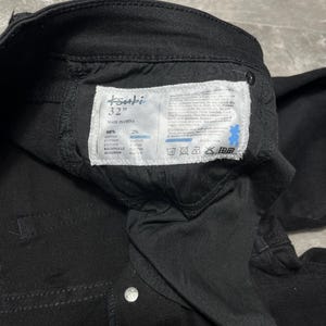 Op de afbeelding: Close-up van een zwarte spijkerbroek met een label met de tekst "tsubi 32" MADE IN CHINA". Het label vermeldt ook de stofsamenstelling als 98% katoen en 2% elastaan. De jeans heeft zilveren knoopsluitingen.
