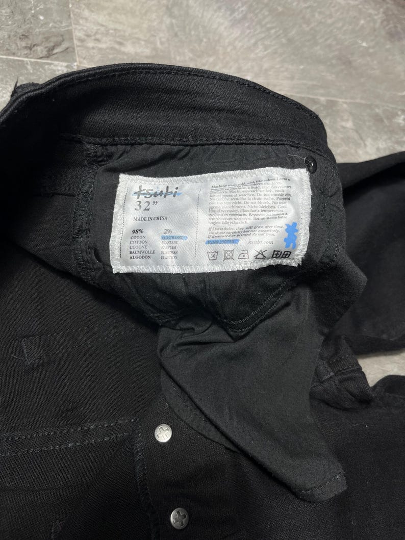 Op de afbeelding: Close-up van een zwarte spijkerbroek met een label met de tekst "tsubi 32" MADE IN CHINA". Het label vermeldt ook de stofsamenstelling als 98% katoen en 2% elastaan. De jeans heeft zilveren knoopsluitingen.