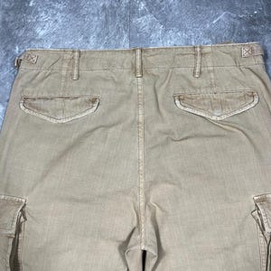 May include: Tan cargo pants featuring a high waistband with belt loops and side adjustment tabs. The back includes two flap pockets with button closures and two large side cargo pockets on the legs, showcasing a durable, woven fabric construction.