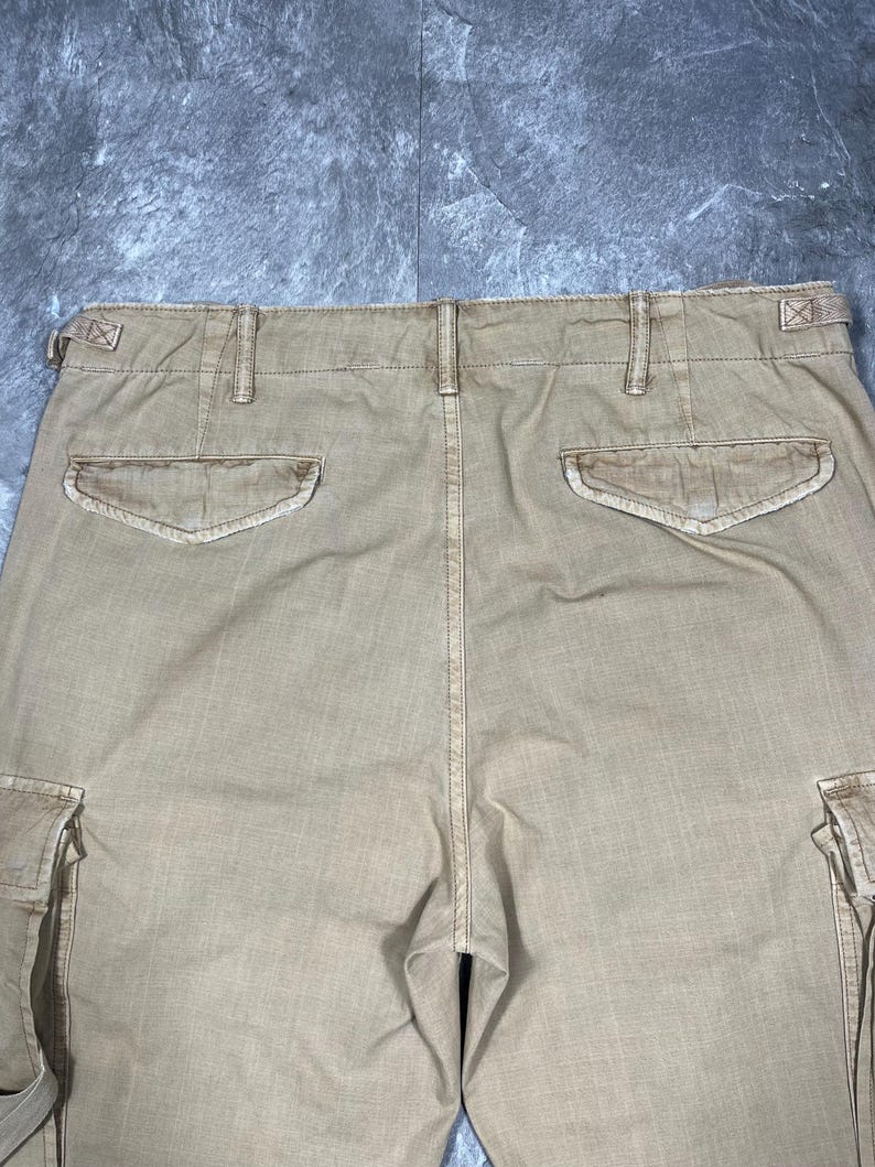 May include: Tan cargo pants featuring a high waistband with belt loops and side adjustment tabs. The back includes two flap pockets with button closures and two large side cargo pockets on the legs, showcasing a durable, woven fabric construction.