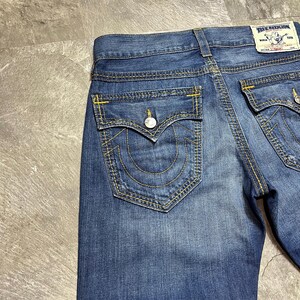 May include: Close-up of blue denim jeans with yellow stitching. The jeans have two back pockets with a curved design and a button closure. A True Religion label is visible on the back.