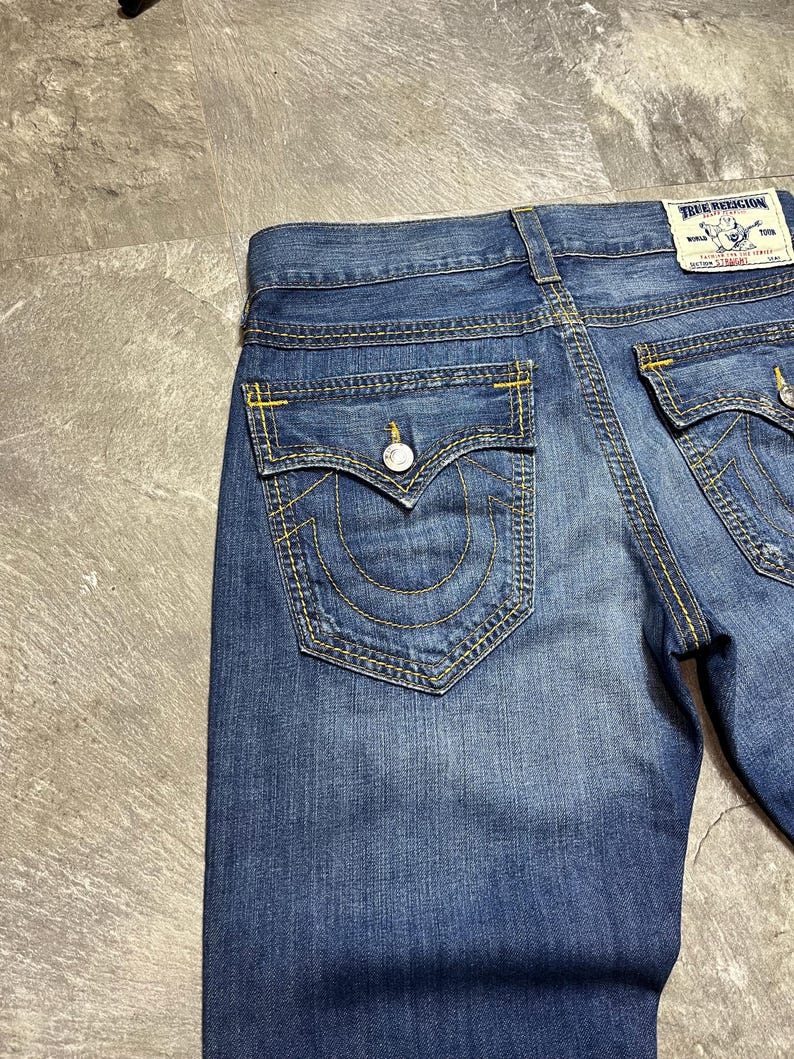May include: Close-up of blue denim jeans with yellow stitching. The jeans have two back pockets with a curved design and a button closure. A True Religion label is visible on the back.