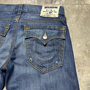 May include: Close-up of blue denim jeans with yellow stitching. The back pocket features a signature horseshoe design and a button closure. A True Religion label is visible.