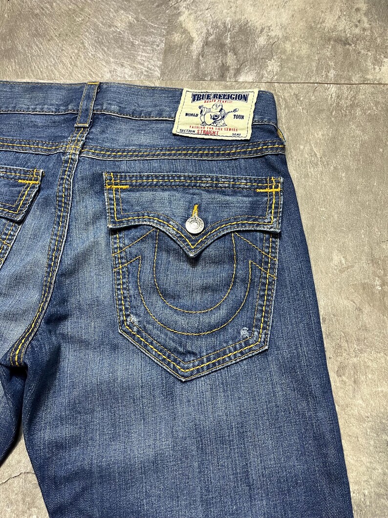May include: Close-up of blue denim jeans with yellow stitching. The back pocket features a signature horseshoe design and a button closure. A True Religion label is visible.