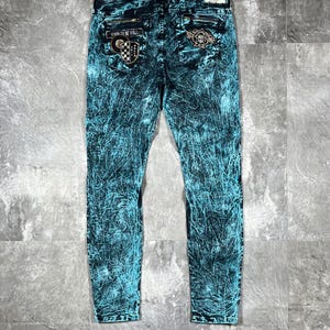 May include: Acid-wash denim jeans in a vibrant teal and black marbled pattern. The back features two rear pockets with zippered accents, a rectangular leather-style patch on the waistband, and two decorative embroidered patches with text.