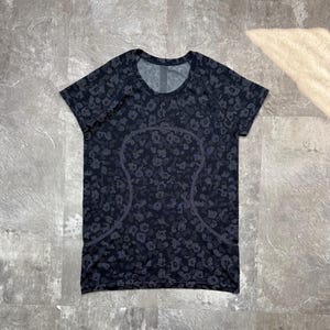 Pode incluir: Uma t-shirt desportiva de manga curta em preto e cinzento escuro com uma construção em malha sem costuras, um padrão de leopardo repetitivo e uma linha de contorno curva distinta na parte frontal do peito e do tronco.
