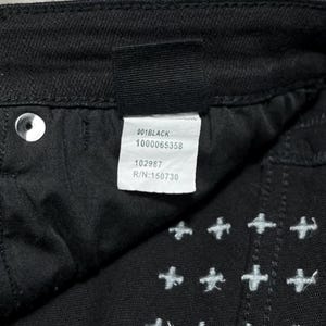 Op de afbeelding: Close-up van een zwarte spijkerbroek met witte kruissteekborduurwerk. Het label luidt "001BLACK", "1000065358", "102987" en "R/N:150730". De broek heeft een knoop en een klinknagel.