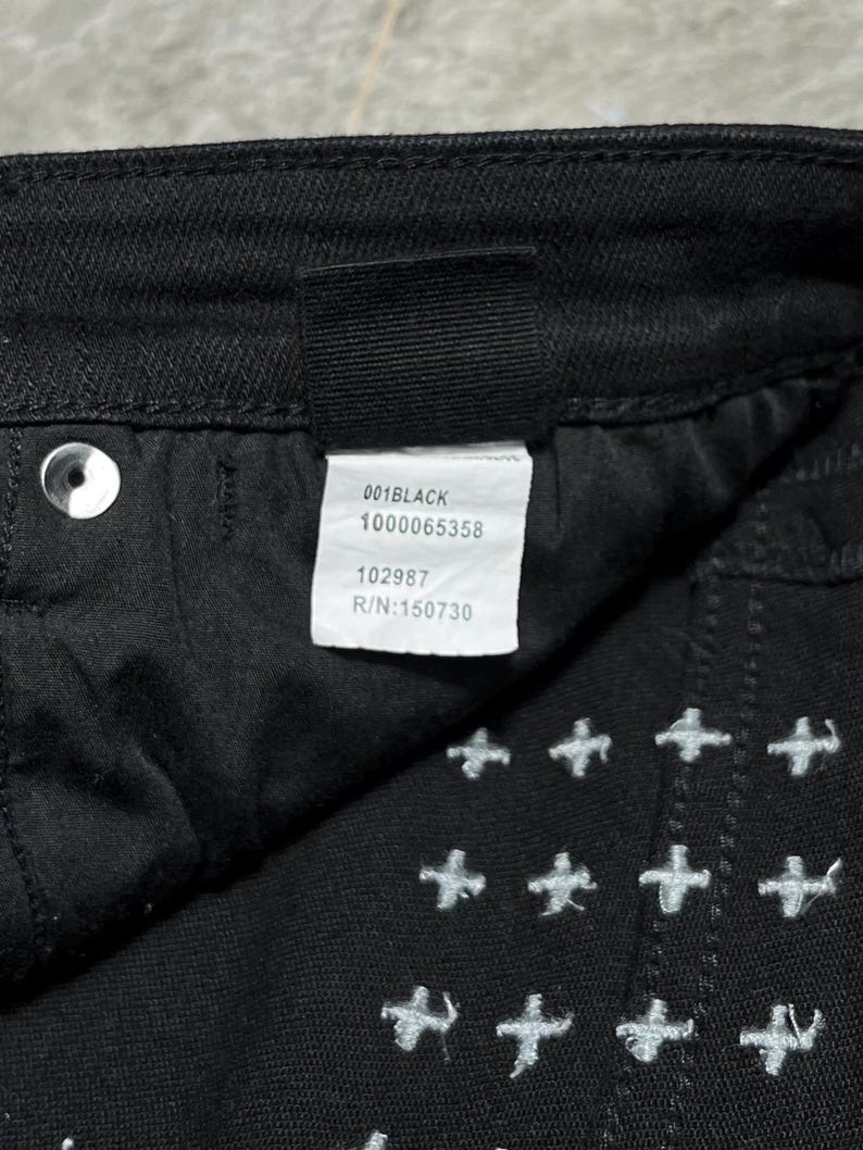 Op de afbeelding: Close-up van een zwarte spijkerbroek met witte kruissteekborduurwerk. Het label luidt "001BLACK", "1000065358", "102987" en "R/N:150730". De broek heeft een knoop en een klinknagel.