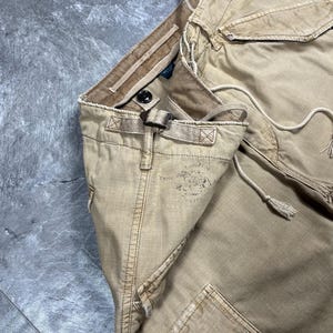 May include: Tan cargo pants featuring a side adjustment buckle, a drawstring waist, and a faded graphic print on the hip. The pants include multiple pockets with flaps, reinforced stitching, and a button closure at the waistband for a secure fit.