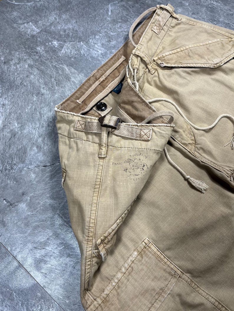 May include: Tan cargo pants featuring a side adjustment buckle, a drawstring waist, and a faded graphic print on the hip. The pants include multiple pockets with flaps, reinforced stitching, and a button closure at the waistband for a secure fit.