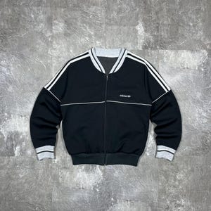 May include: Black zip-up jacket with white stripes on the sleeves and shoulders. The jacket has a gray collar and cuffs. The Adidas logo is on the chest.