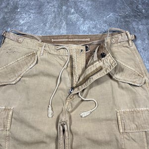May include: Tan cargo pants featuring a front zipper closure, a drawstring waist with tasseled ends, and two large side cargo pockets. The waistband includes belt loops and side adjustment straps. The fabric has a visible textured weave.