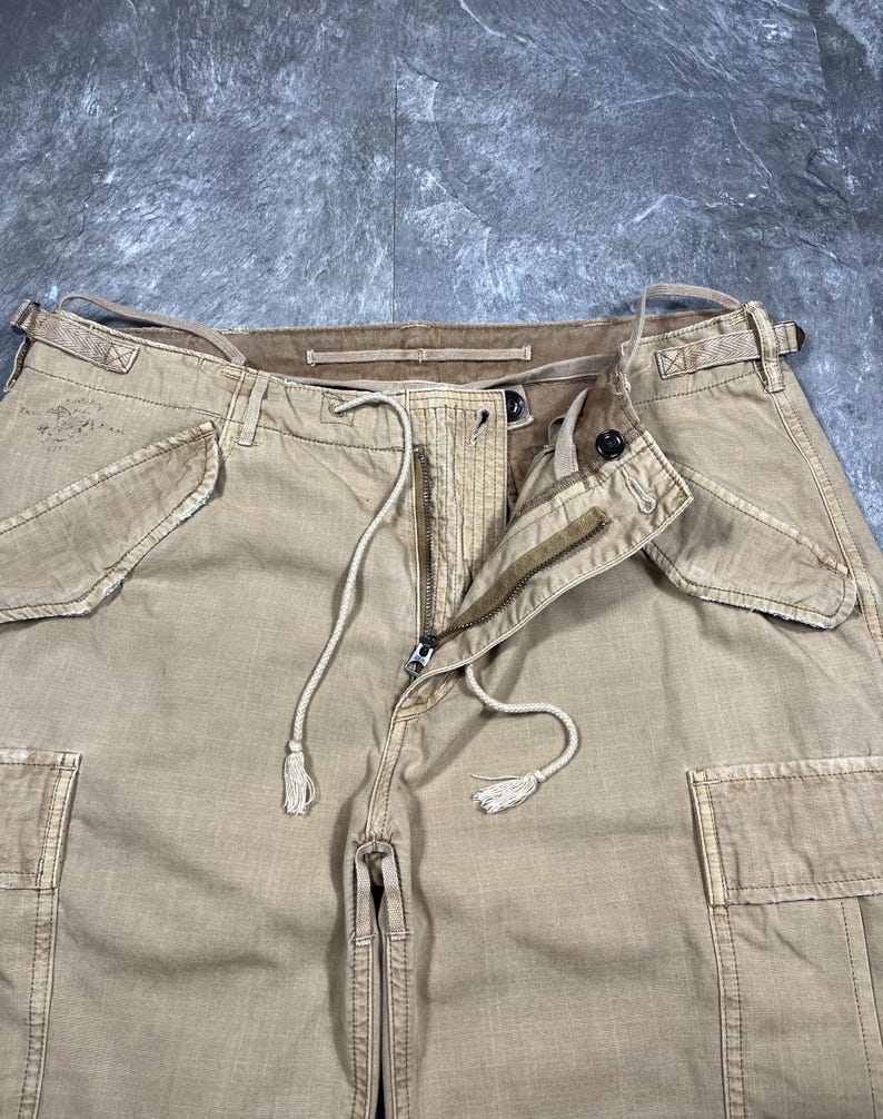 May include: Tan cargo pants featuring a front zipper closure, a drawstring waist with tasseled ends, and two large side cargo pockets. The waistband includes belt loops and side adjustment straps. The fabric has a visible textured weave.