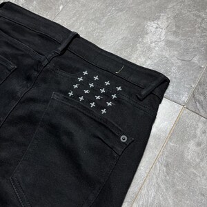 Op de afbeelding: Close-up van een zwarte denim jeans. De achterzak heeft een patroon van kleine, wit geborduurde kruisjes. De jeans heeft een klassiek ontwerp met vijf zakken en een knoopsluiting. De stof lijkt een medium-gewicht denim te zijn.