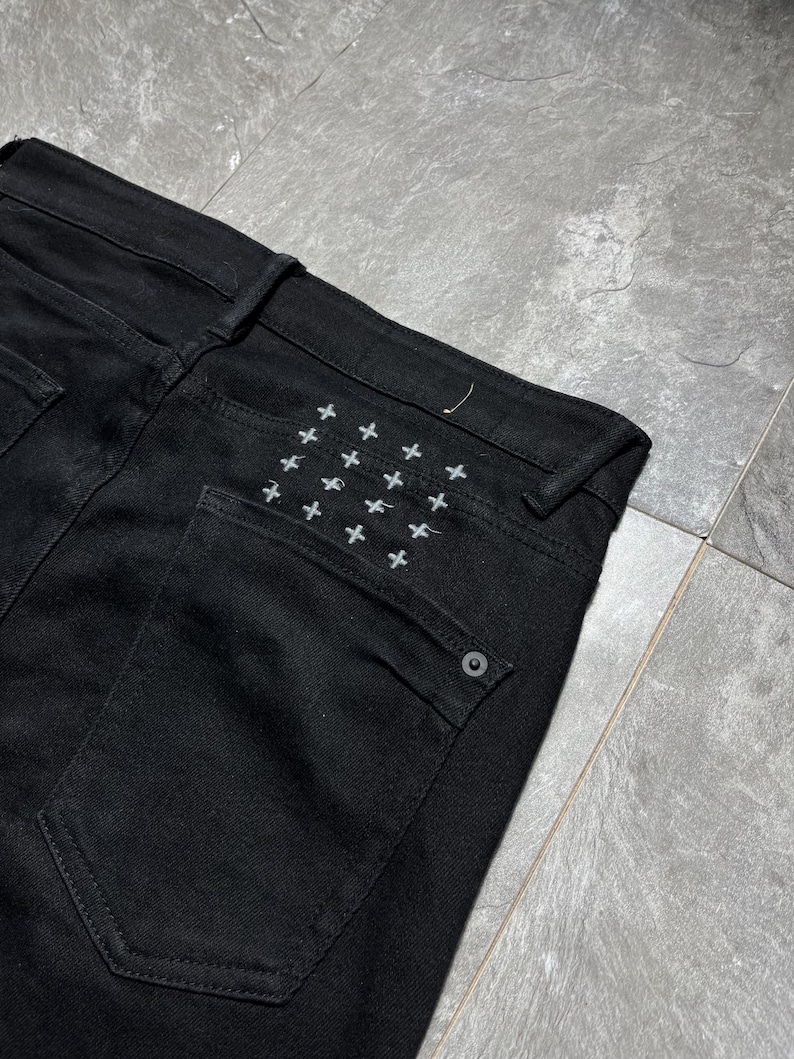 Op de afbeelding: Close-up van een zwarte denim jeans. De achterzak heeft een patroon van kleine, wit geborduurde kruisjes. De jeans heeft een klassiek ontwerp met vijf zakken en een knoopsluiting. De stof lijkt een medium-gewicht denim te zijn.