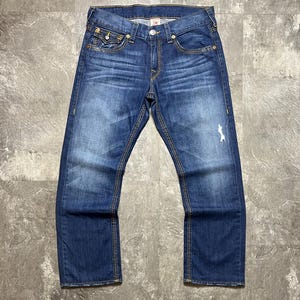May include: Blue denim jeans with a faded wash and distressed details. The jeans feature classic five-pocket styling, a button and zipper closure, and yellow stitching. The jeans are laid flat on a gray surface.