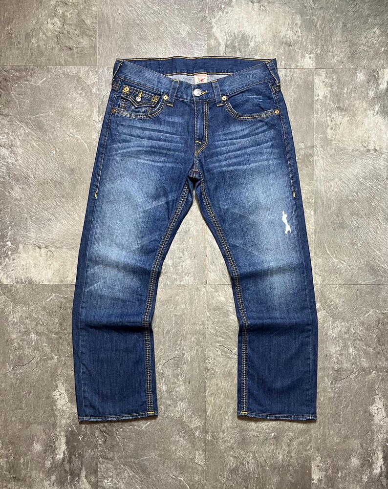 May include: Blue denim jeans with a faded wash and distressed details. The jeans feature classic five-pocket styling, a button and zipper closure, and yellow stitching. The jeans are laid flat on a gray surface.