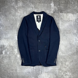May include: A dark navy blue blazer featuring a notched lapel, two front buttons, and three smaller buttons on each cuff. The interior lining is white with a black G-Star Raw Correct label at the neck. It includes two front pockets and one chest pocket.