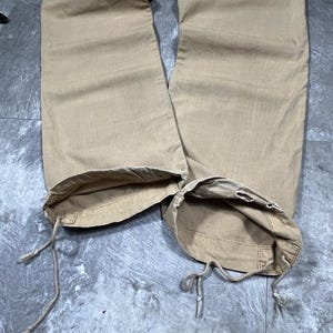 May include: Tan cargo pants featuring wide legs and a straight cut. The hem of each pant leg includes an integrated drawstring cord for adjustment, allowing the opening to be cinched. The fabric has a visible weave texture throughout the garment.