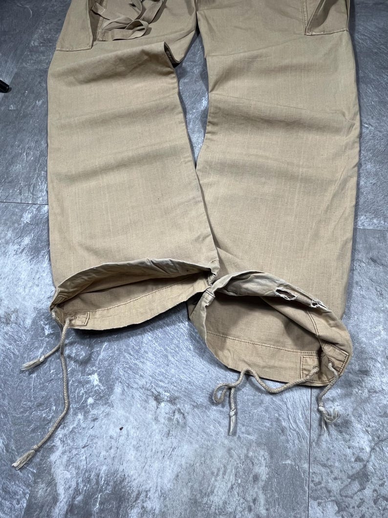 May include: Tan cargo pants featuring wide legs and a straight cut. The hem of each pant leg includes an integrated drawstring cord for adjustment, allowing the opening to be cinched. The fabric has a visible weave texture throughout the garment.