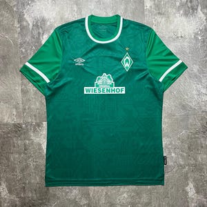 Könnte beinhalten: Grünes Fußballtrikot mit weißen Akzenten an Kragen und Ärmeln. Das Trikot zeigt das Umbro-Logo, ein Teamwappen und das Logo "WIESENHOF". Das Trikot hat ein strukturiertes Muster und einen kleinen Stern über dem Teamwappen.